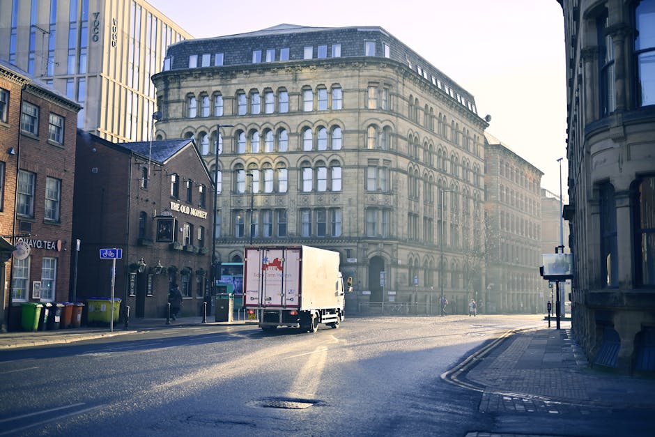 Image depicts a city street scene during daylight hours, with a white moving truck positioned on the road, preparing for a house relocation. The truck displays a red logo and is situated near a tall, 