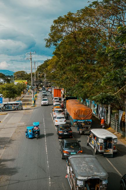 A busy street scene showing multiple vehicles including cars, vans, and a tuk-tuk driving along a road with an adjacent pavement. On the right side, there are tall trees with dense green foliage, casting shade over the sidewalk and storefronts. Parked and moving vehicles include a large truck with an orange tarpaulin covering its load, a black car, and a small tuk-tuk with a blue roof. The street is lined with utility poles and overhead wiring, with cloudy skies overhead. The scene captures typical urban moving logistics, where furniture transport and home relocation activities involving loading and unloading could be taking place, supported by a professional removals company like Archway Removals.